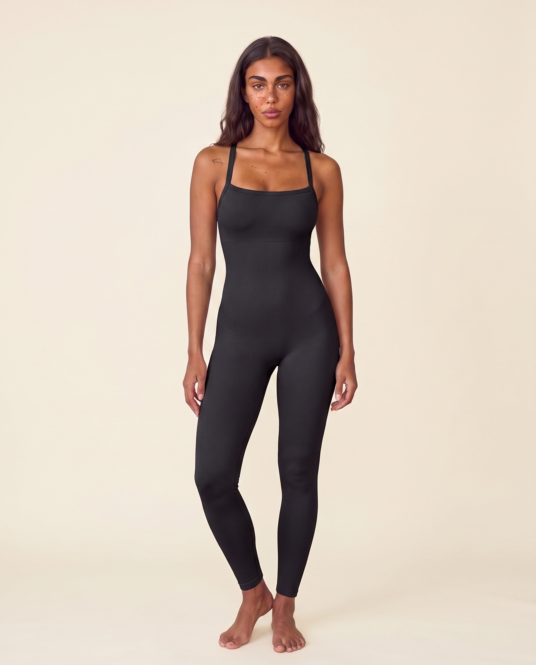 A woman stands facing forward in a black full-length Saturn Squeeze shaping jumpsuit against a warm cream studio backdrop.