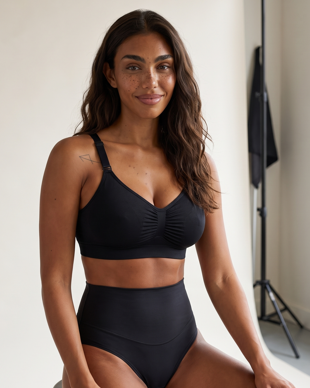 No-Show Tummy Control Thong in Black — seated on a studio stool.